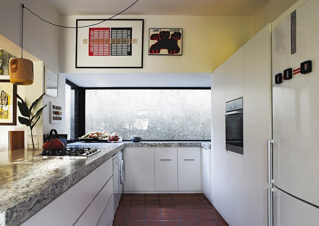 Modern, white fitted kitchen with solid stone worksurface and wide window with view of wall