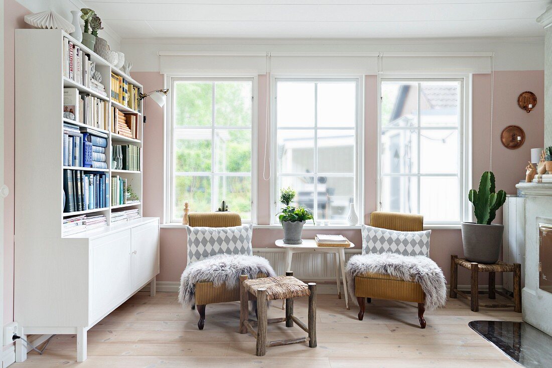 Patterned scatter cushions and fur blankets on two armchairs flanking side table below window and next to dresser with bookshelves