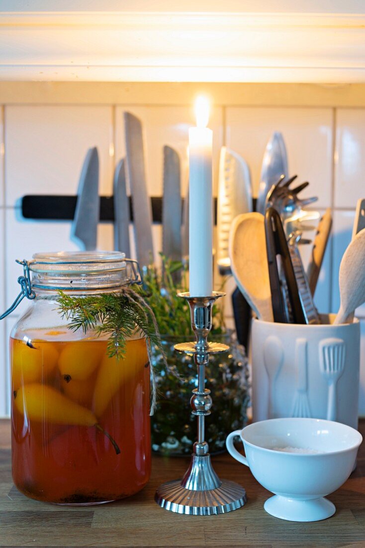 Brennende Kerze und Einmachglas mit eingelegten Birnen und Tannenzweig-Deko vor Küchenutensilien