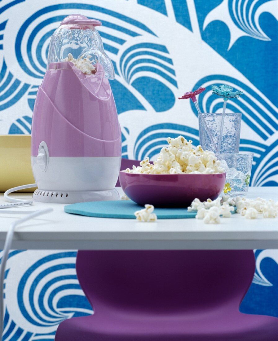 Kultige Popcornmaschine in Lila auf Esstisch, im Hintergrund Stoffbahn mit blau-weißem Wellenmotiv