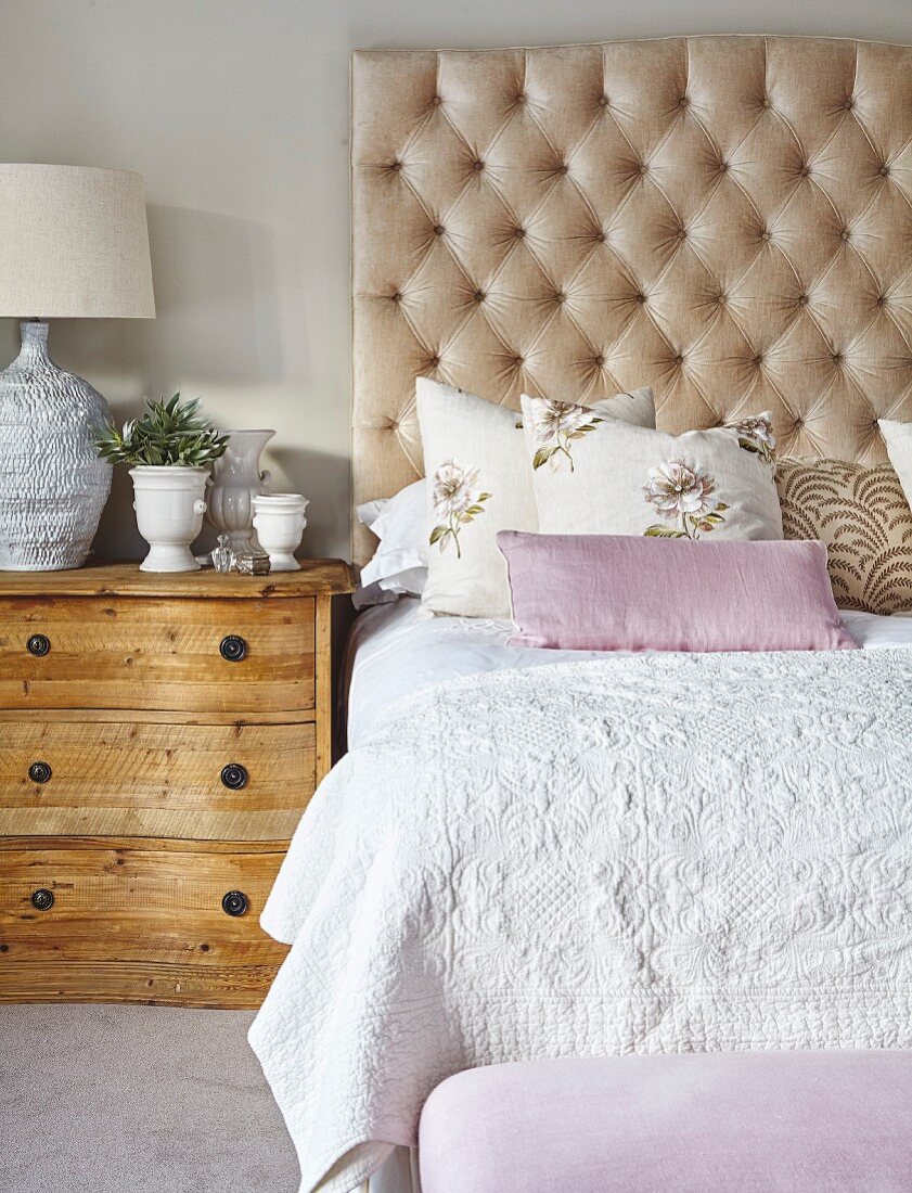 Doppelbett mit kapitoniertem Kopfteil, Tagesdecke mit Prägemuster und floralen Zierkissen, daneben rustikale Landhauskommode