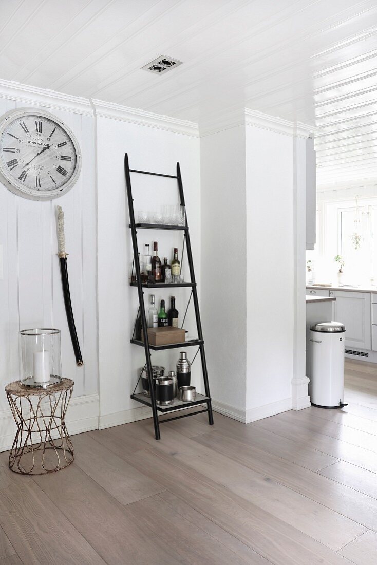 Ladder shelves used as drinks cabinet next to candle lantern on side table below station clock in open-plan interior