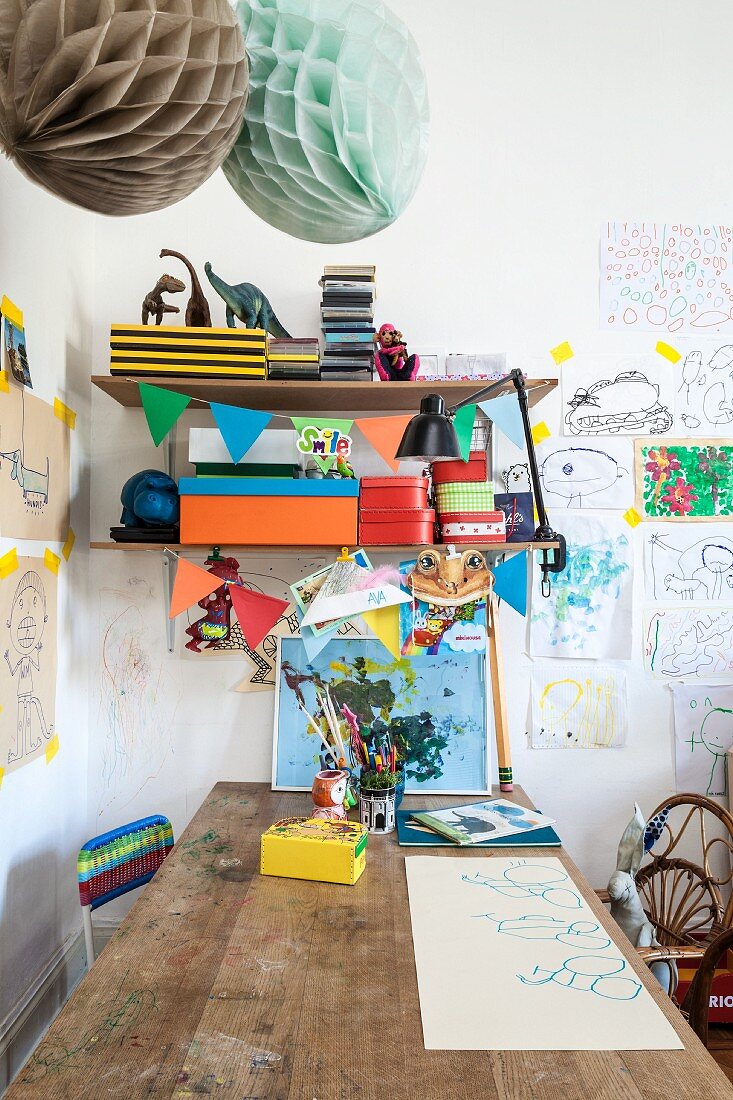 Wooden table below colourful boxes on bracket shelves and children's drawings on wall