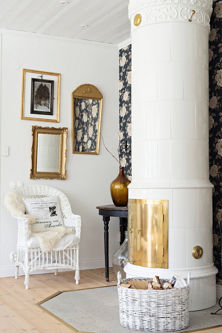 Gilt-framed pictures and mirror above white wicker armchair behind Swedish tiled stove