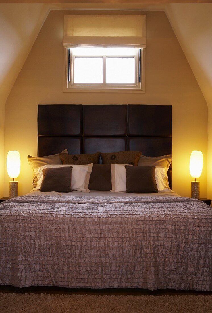 Bed with scatter cushions against dark brown leather headboard flanked by artistic lamps in attic bedroom