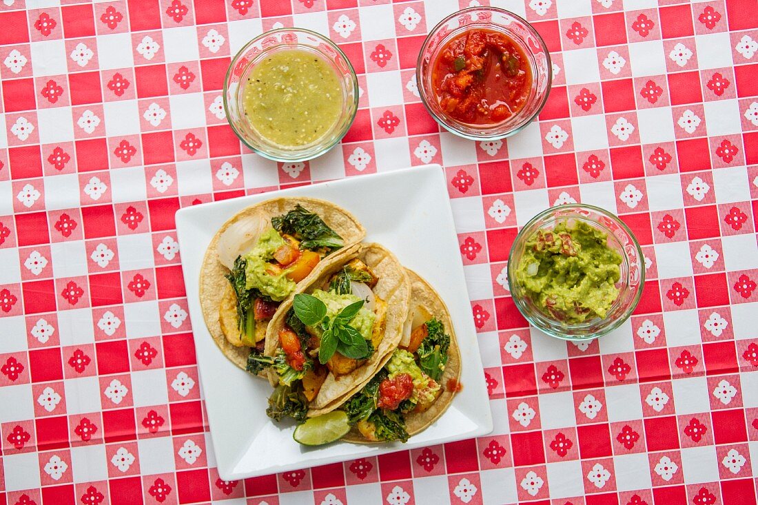 Vegan tacos with kale, guacamole, salsa, and salsa verde on a red-and-white checked tablecloth