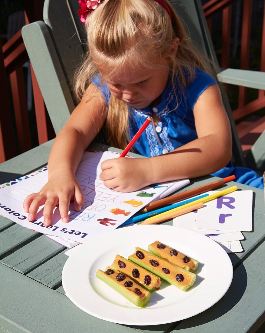 A little girl doing homework with a vegetable snack on a plate next to her