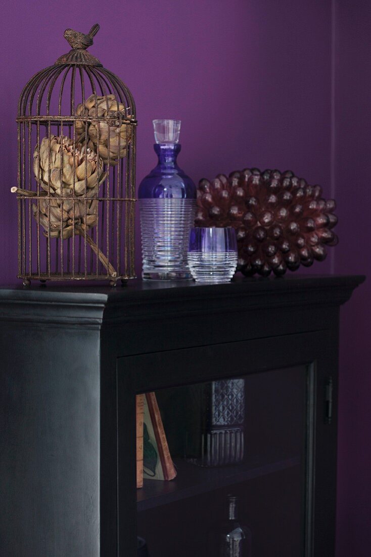 Vintage bird cage on antique, glass-fronted cabinet against aubergine wall