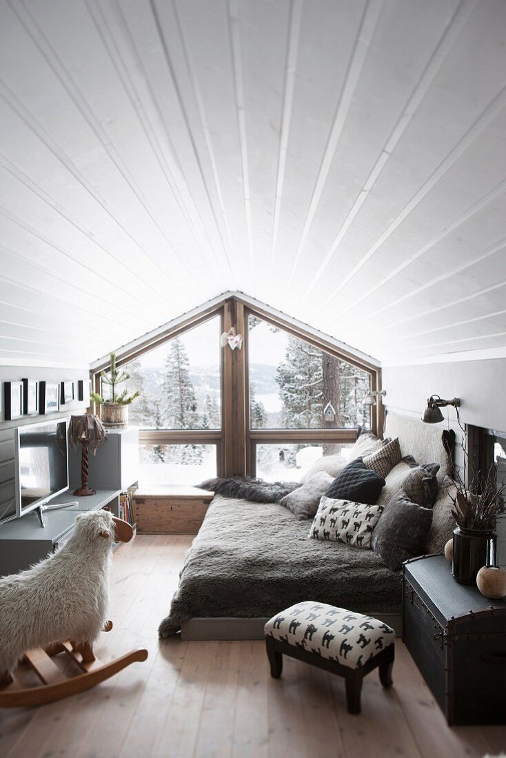 Gemütliches Zimmer mit weisser Holzverschalung unter dem Dach, Schaukelschaf vor gemütlichem Bettlager neben Fensterfront in Giebelwand