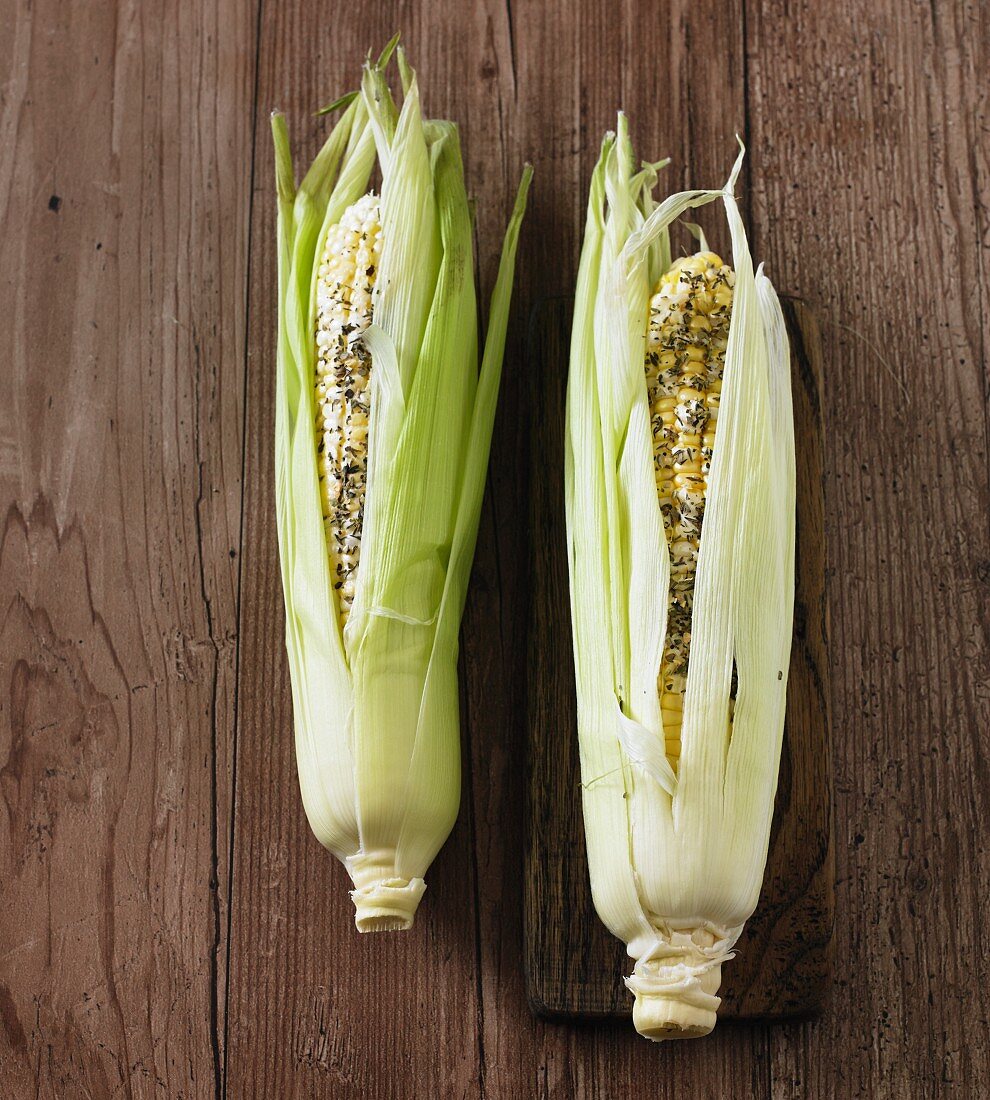 Two fresh corn cobs with butter and herbs, ready to cook
