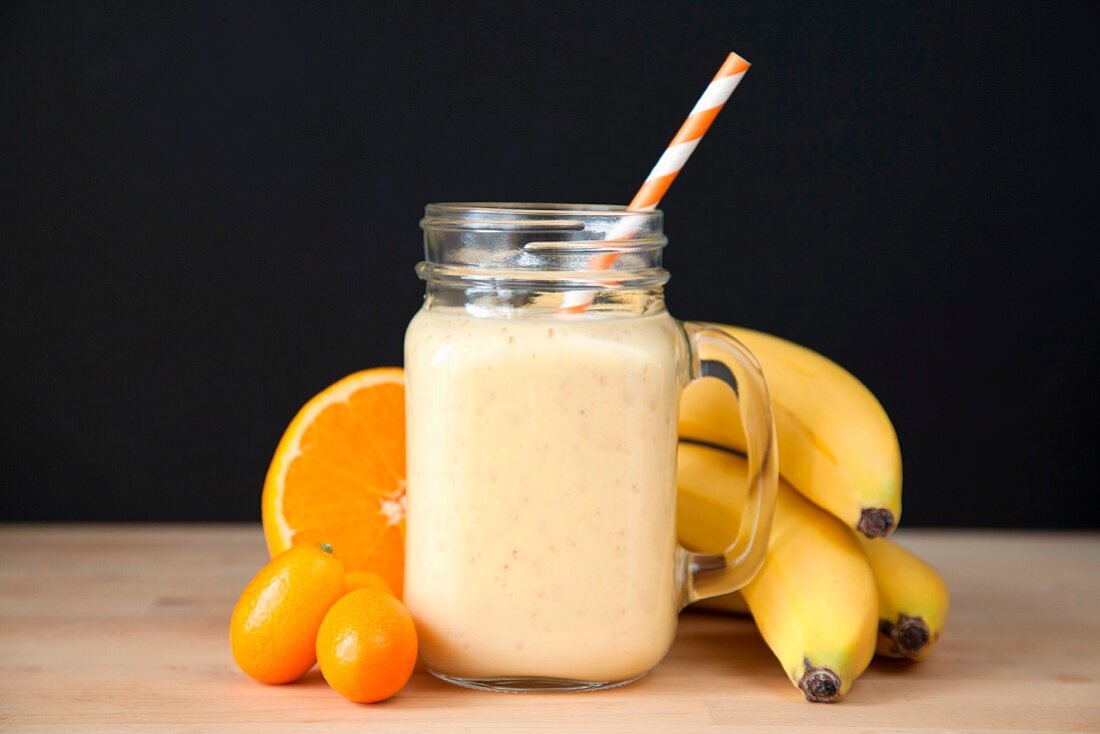 A banana, orange and kumquat smoothie in a glass jug with ingredients