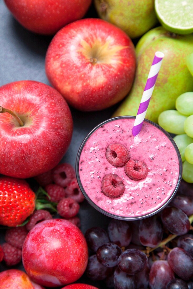 A raspberry smoothie in a glass surrounded by fresh fruit (seen from above)