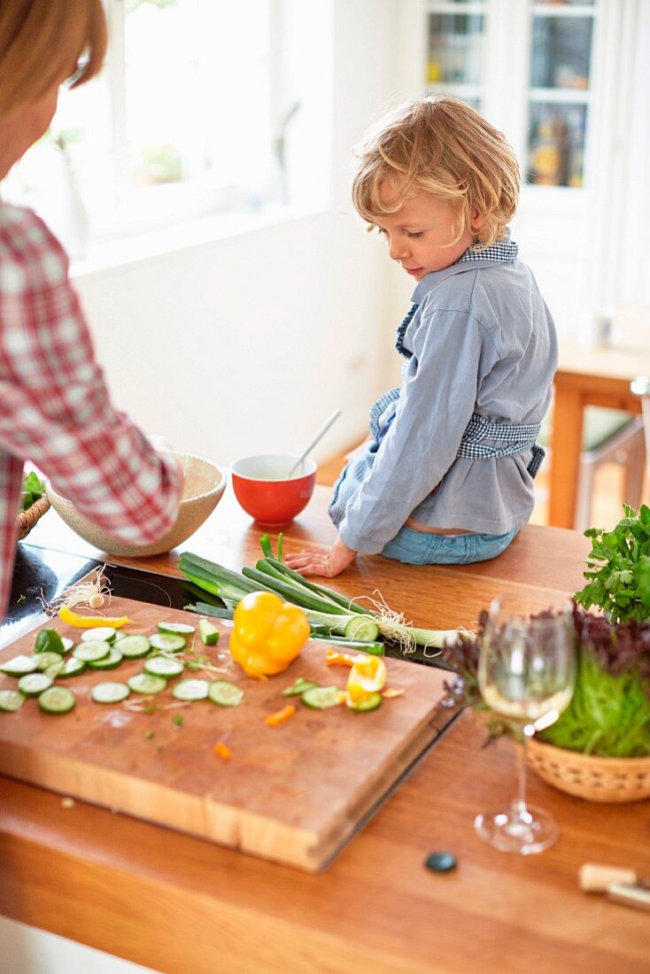 Kind sitzt auf Küchentisch und schaut Mutter beim Zubereiten von Salat zu