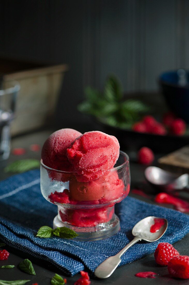 A glass of raspberry sorbet on a blue napkin with a spoon, and fresh raspberries