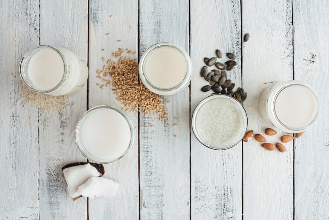 Various types of vegan milk in glasses next to almonds, rice, oats, pumpkin seeds and coconut