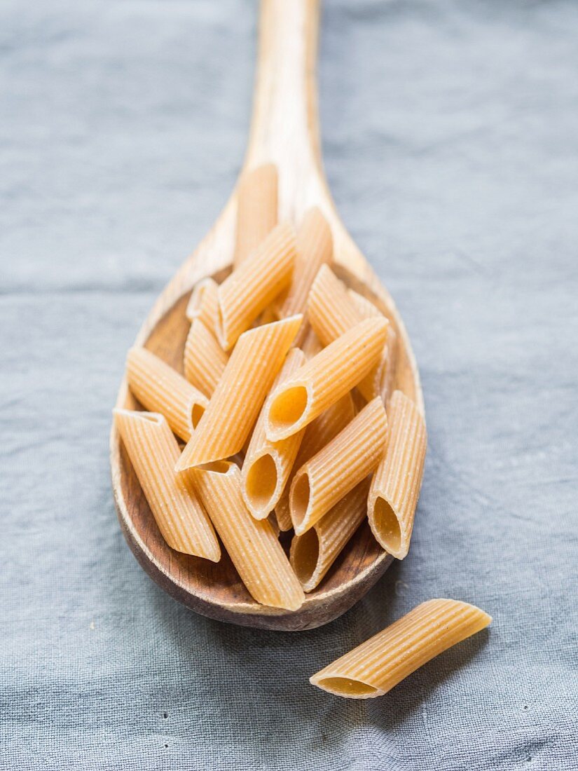 Wholemeal penne pasta on a wooden spoon on a light-blue tablecloth