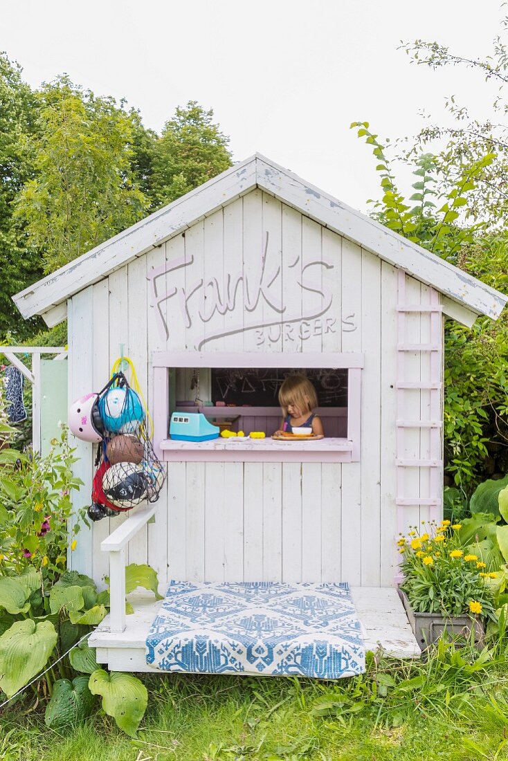 Little girl playing in wooden play house with window in gable end in summery garden