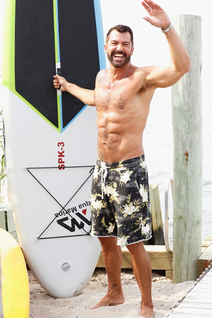 A dark-haired man on a beach wearing bathing shorts with a surfboard