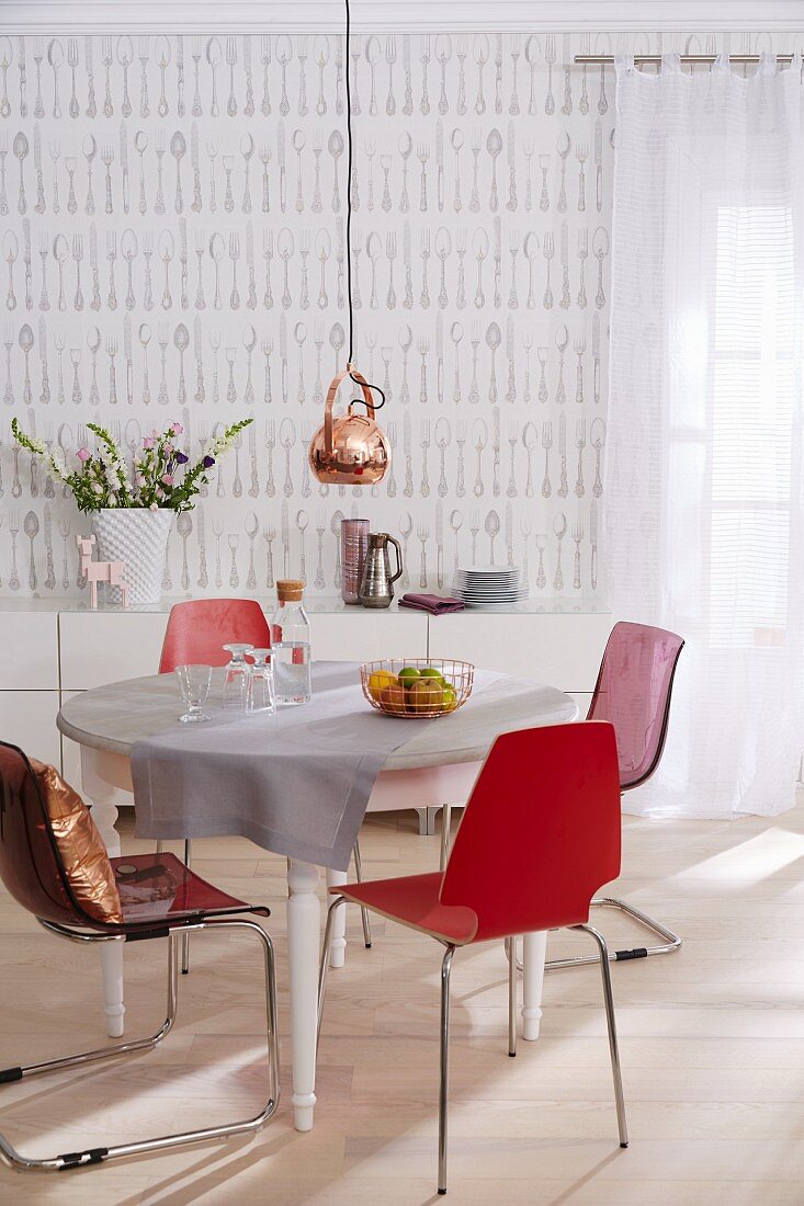 A pendant lamp with a copper shade above a round table with red chairs in a room with cutlery patterned wallpaper