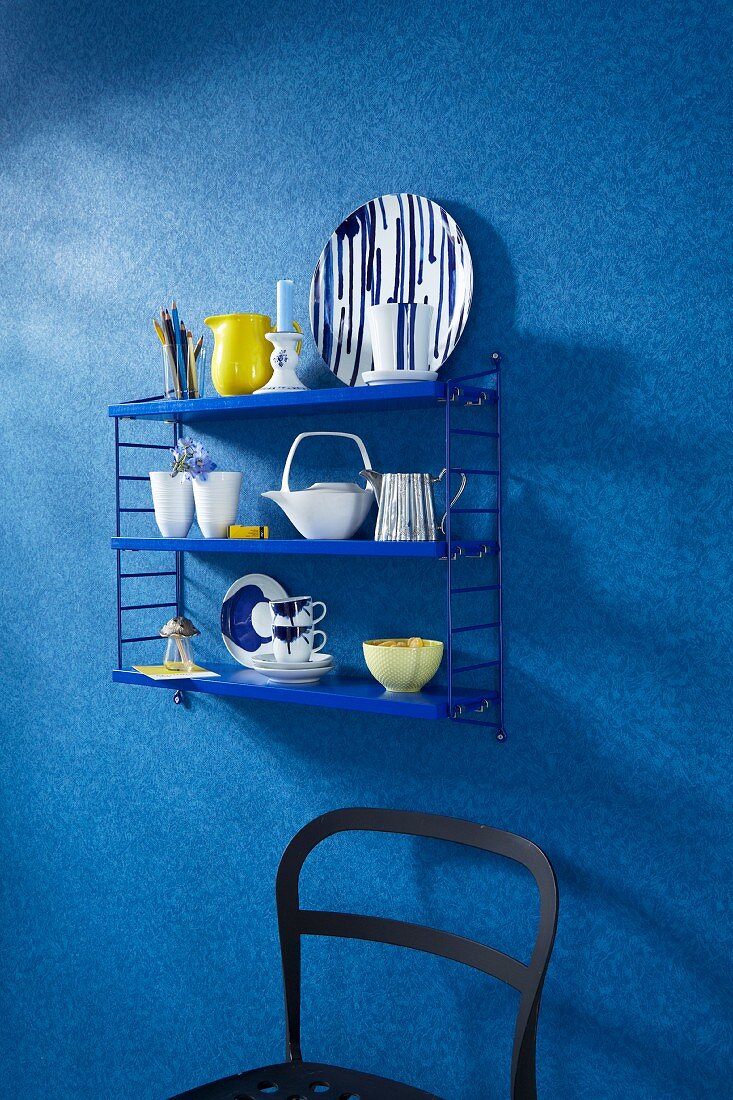 Crockery on a blue shelf on a wall with a blue faux uni patterned wallpaper above a partially visible black chair
