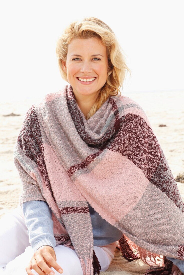 A blonde woman wearing a woollen shawl on a beach