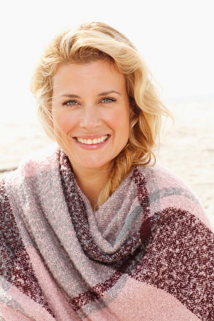 A blonde woman wearing a woollen shawl on a beach