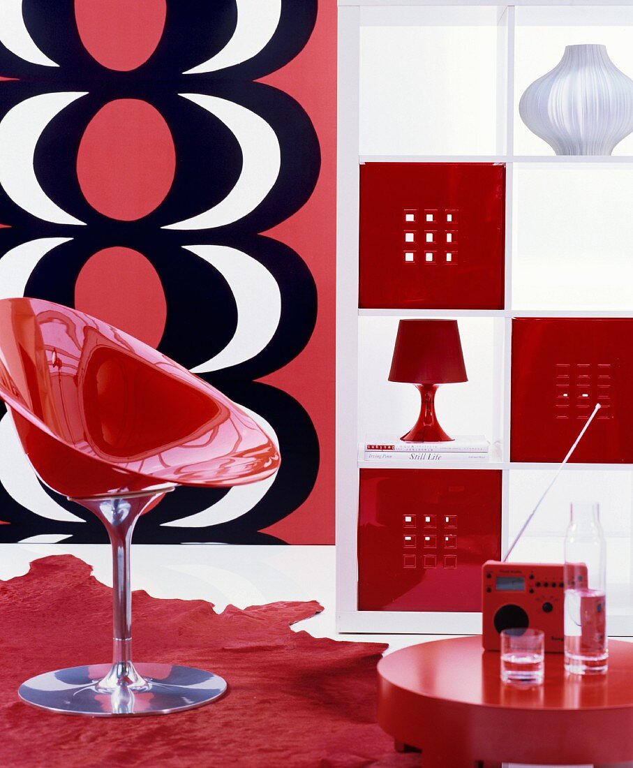 Wohnbereich mit rotem Schalenstuhl, offenem weißem Regal und Wanddekoration mit rot-weiß-schwarzem Muster im Retrostil