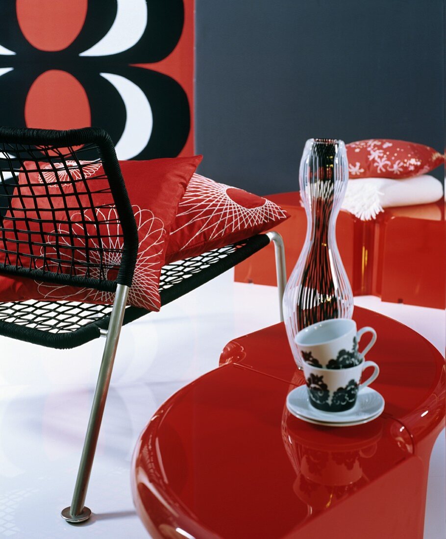 Schwarzer Geflechtstuhl, roter Beistelltisch und Tassen, Wanddekoration mit rot-weiß-schwarzem Muster im Retrostil