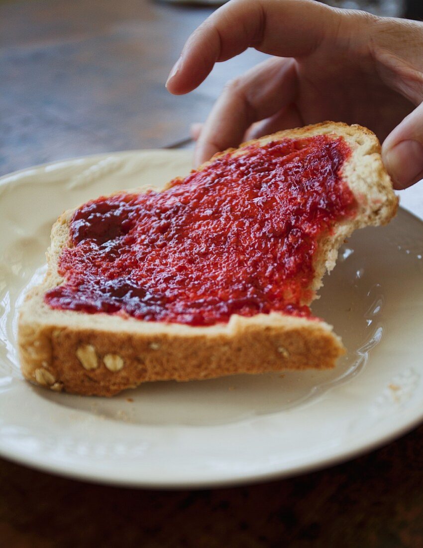 Frauenhand hält angebissene Toastscheibe mit Erdbeermarmelade