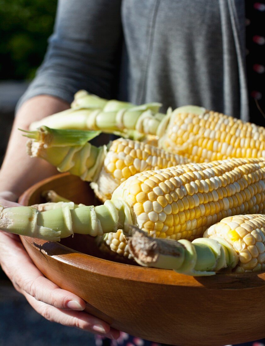 A woman holding a wooden bowl of corn cobs