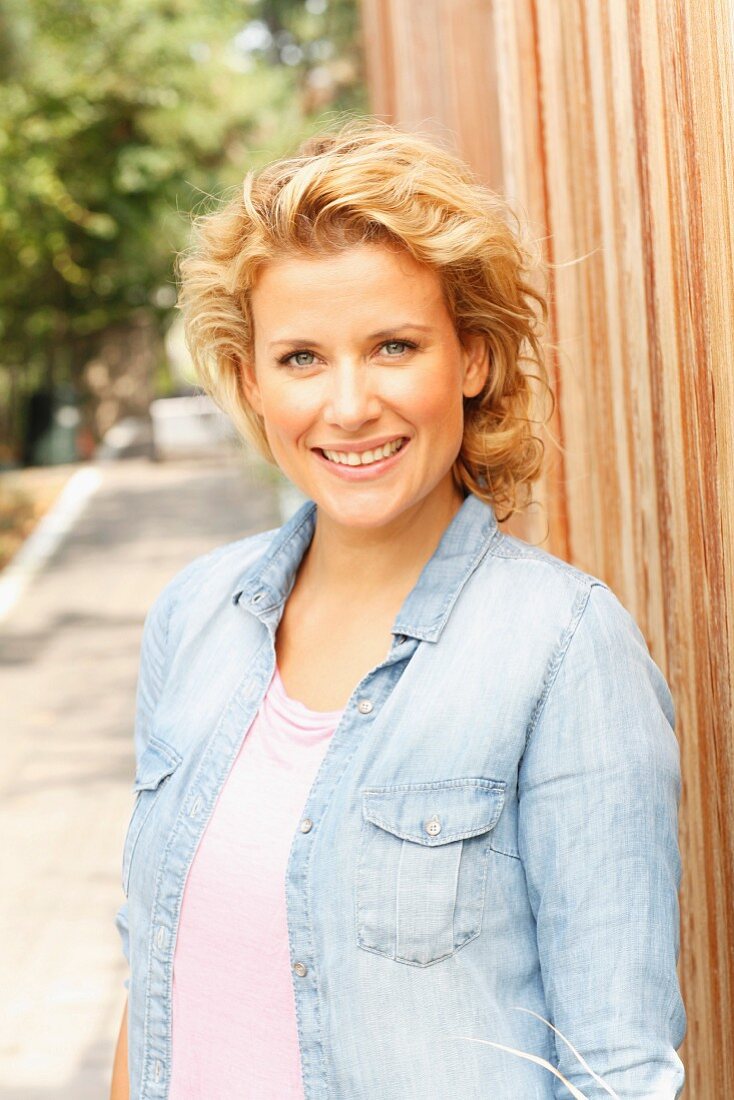 A blonde woman wearing a denim shirt leaning on a fence