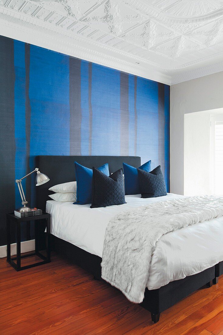 Elegantes Schlafzimmer mit schwarzem Doppelbett und drapierten Kissen, Tapete mit blauschwarzem Streifenmuster und weiße Stuckdecke