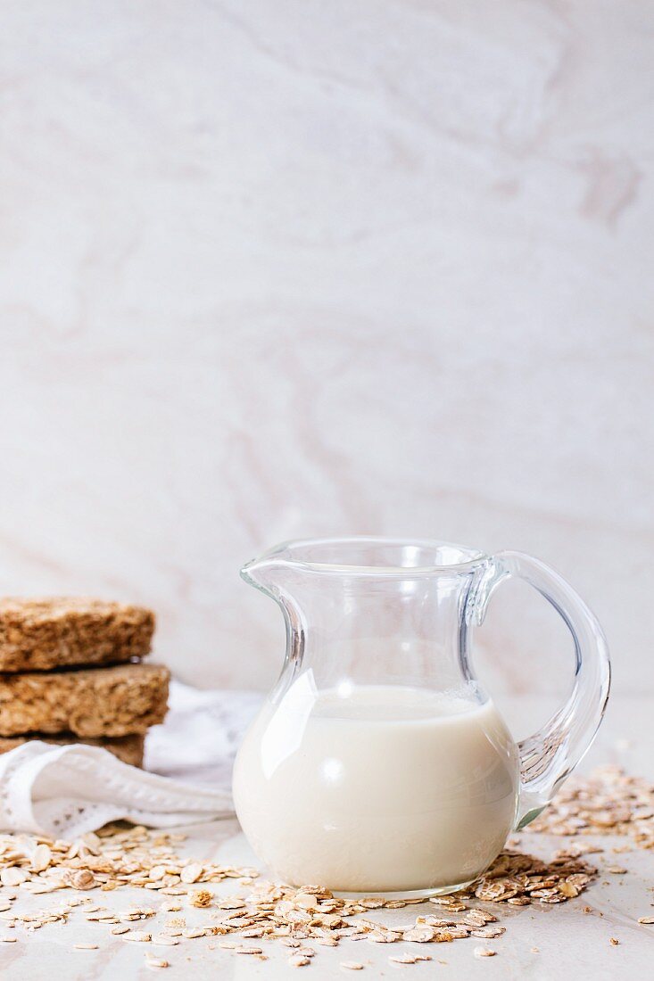 A jug of oat milk, oats and crispbread on a white marble surface