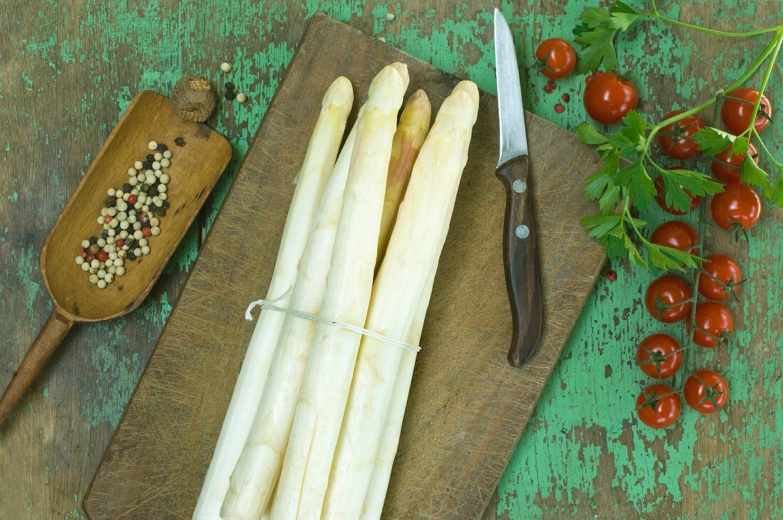 A bundle of fresh white asparagus, tomatoes and parsley on a wooden table, peppercorns in a mortar and a knife