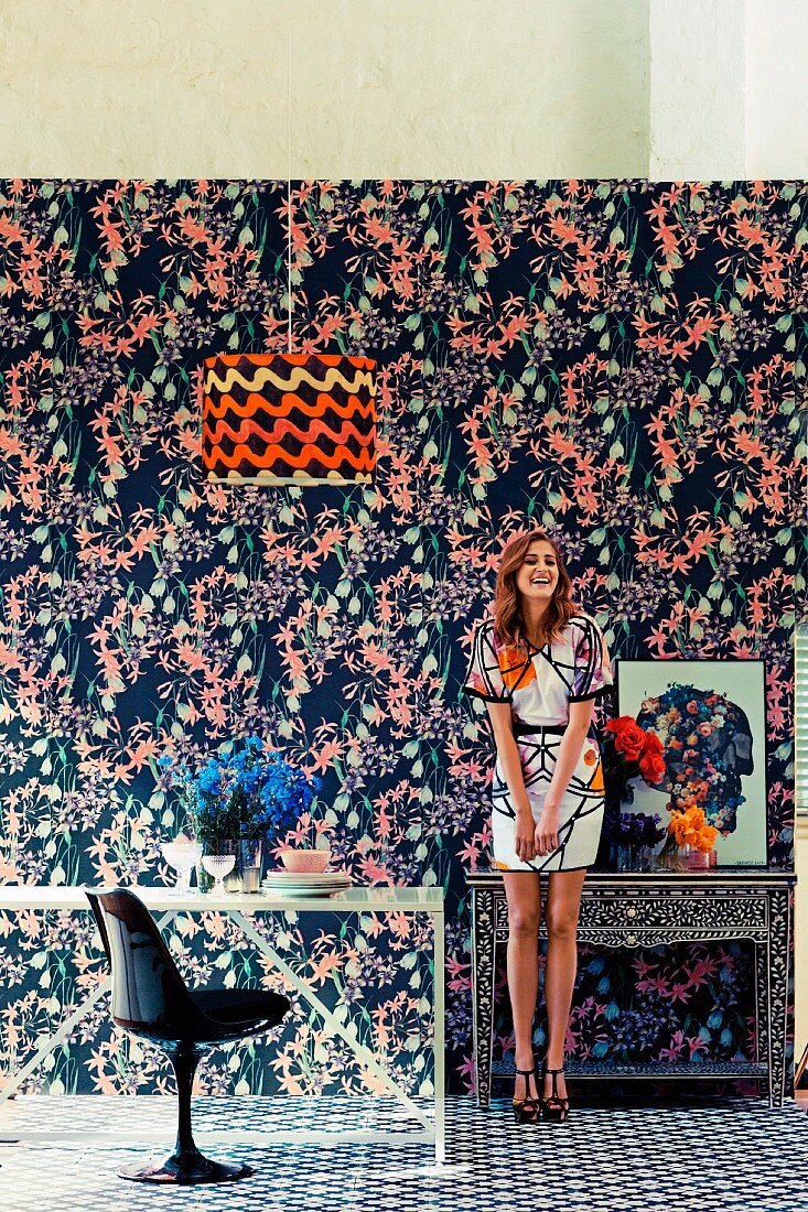Junge Frau in Zimmer mit verschiedenen Mustern an Tapete, Teppich, Lampenschirm und Kleid