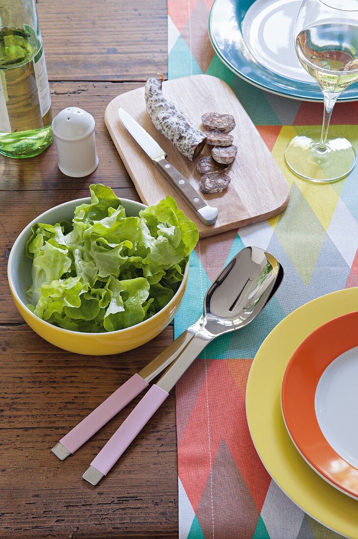 Gedeckter Tisch mit bunten Accessoires, Salami, Salat und Weißwein