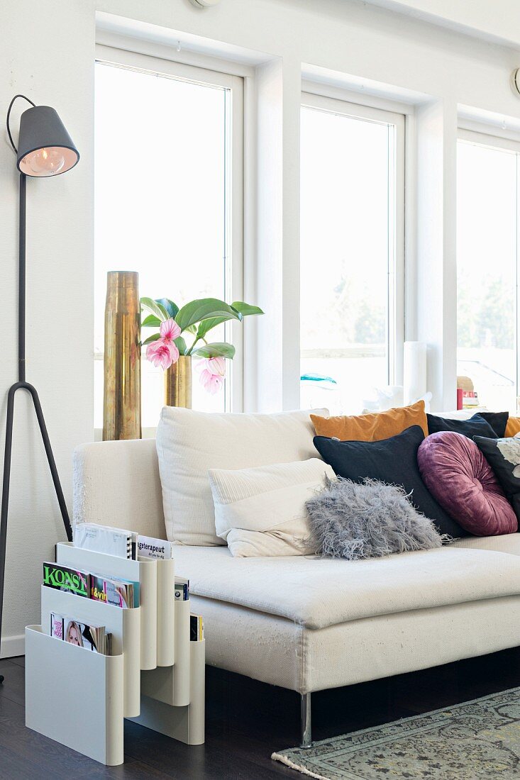 Magazine rack and designer standard lamp next to many cushions on pale couch below window