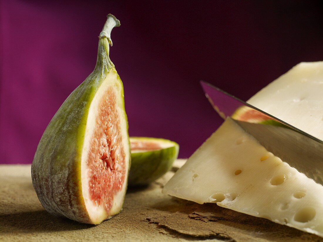 A halved fig next to a slice of cheese with a knife on a natural stone platter