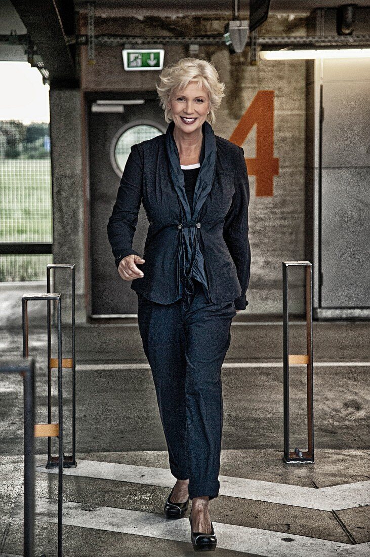 A blonde woman wearing a black cord trousers and a jacket in a multi-storey car park