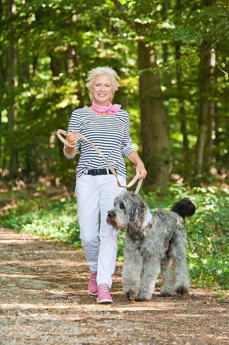 A blonde woman wearing a striped top, white trousers and pink shoes in a forest with a dog