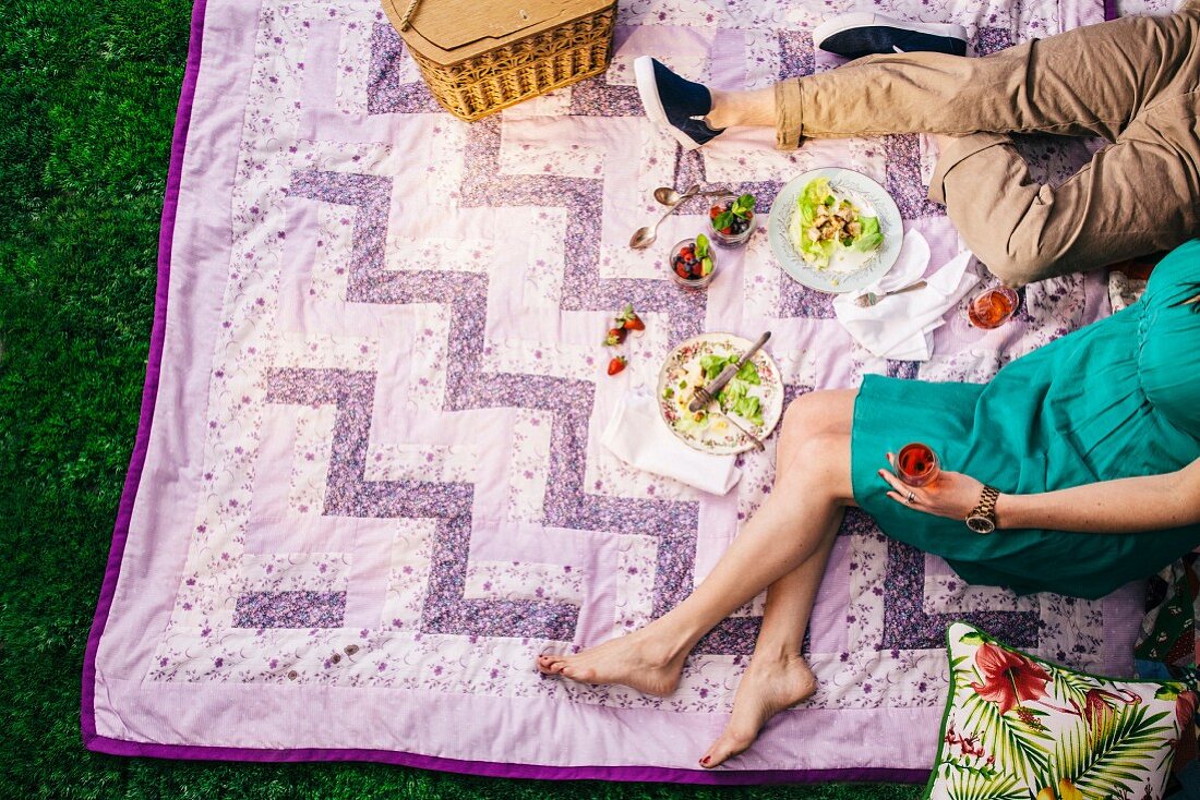 A couple sitting on a patchwork blanket with glasses of red wine and a picnic basket