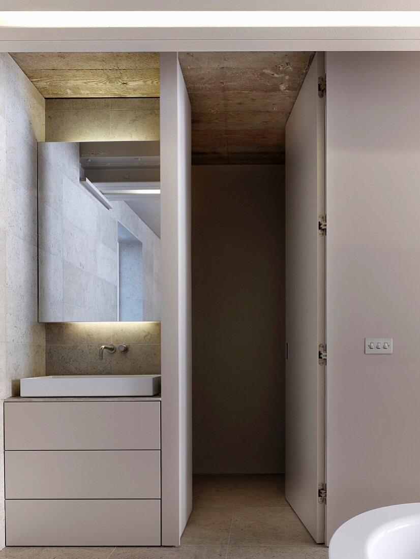 Washstand with drawers in base unit and mirror built into niche with indirect lighting; open door leading to toilet area