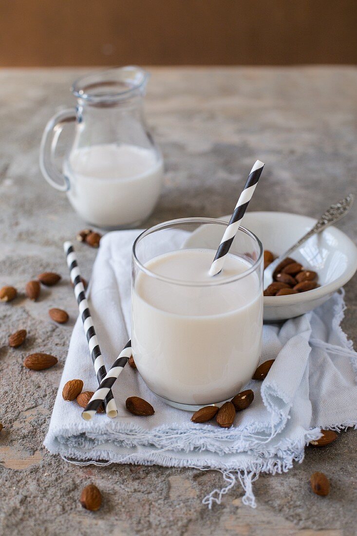 Almond milk in glass with a black and white striped straw, a jug of milk and scattered almonds