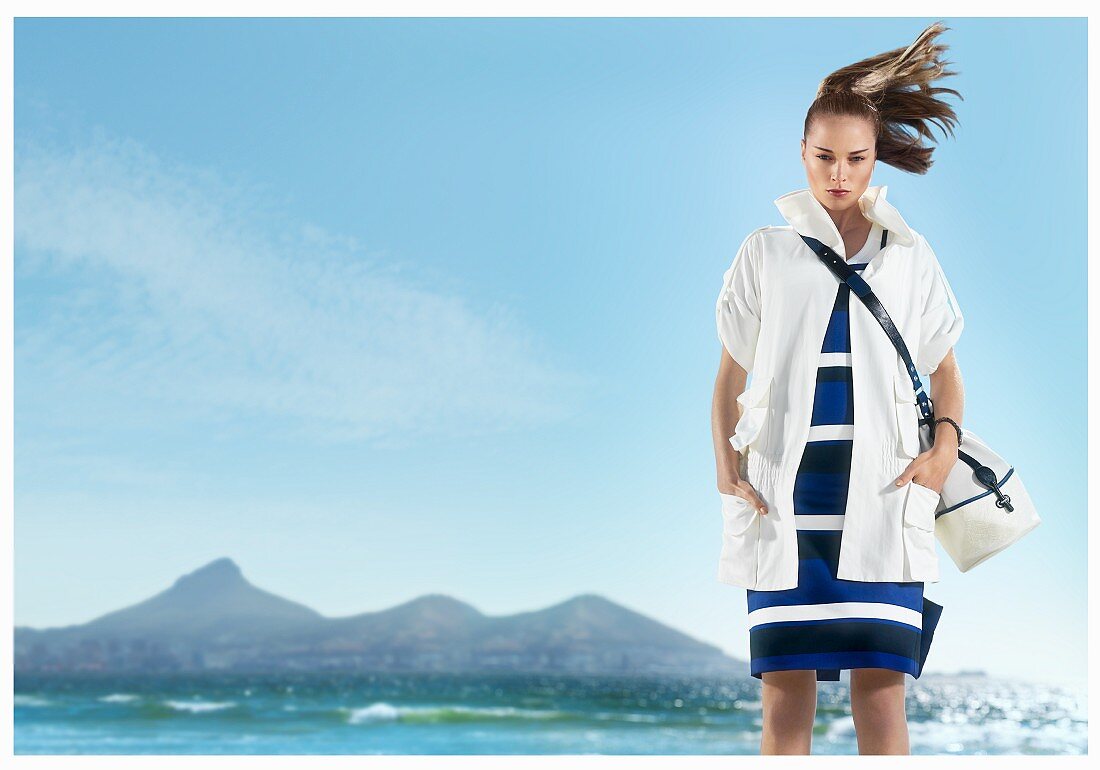 A woman on a beach wearing a blue, white and black striped dress and white jacket