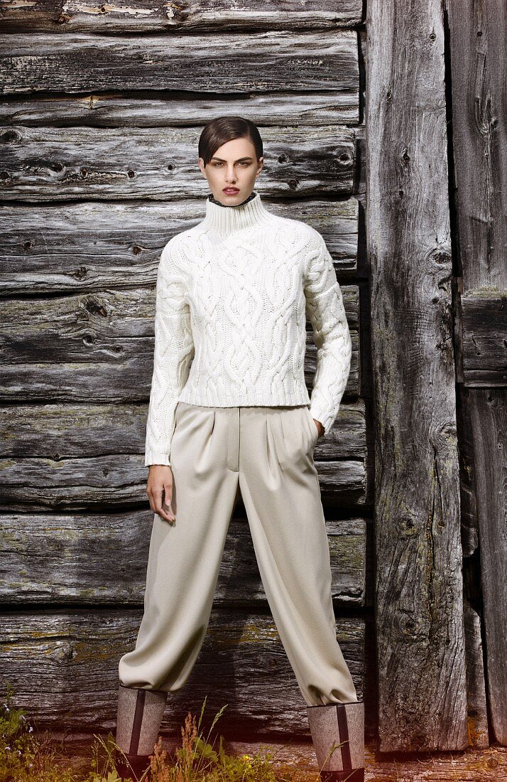 A short-haired woman wearing a cable-knit turtleneck jumper and wide-cut pleated trousers standing in front of a wooden hut