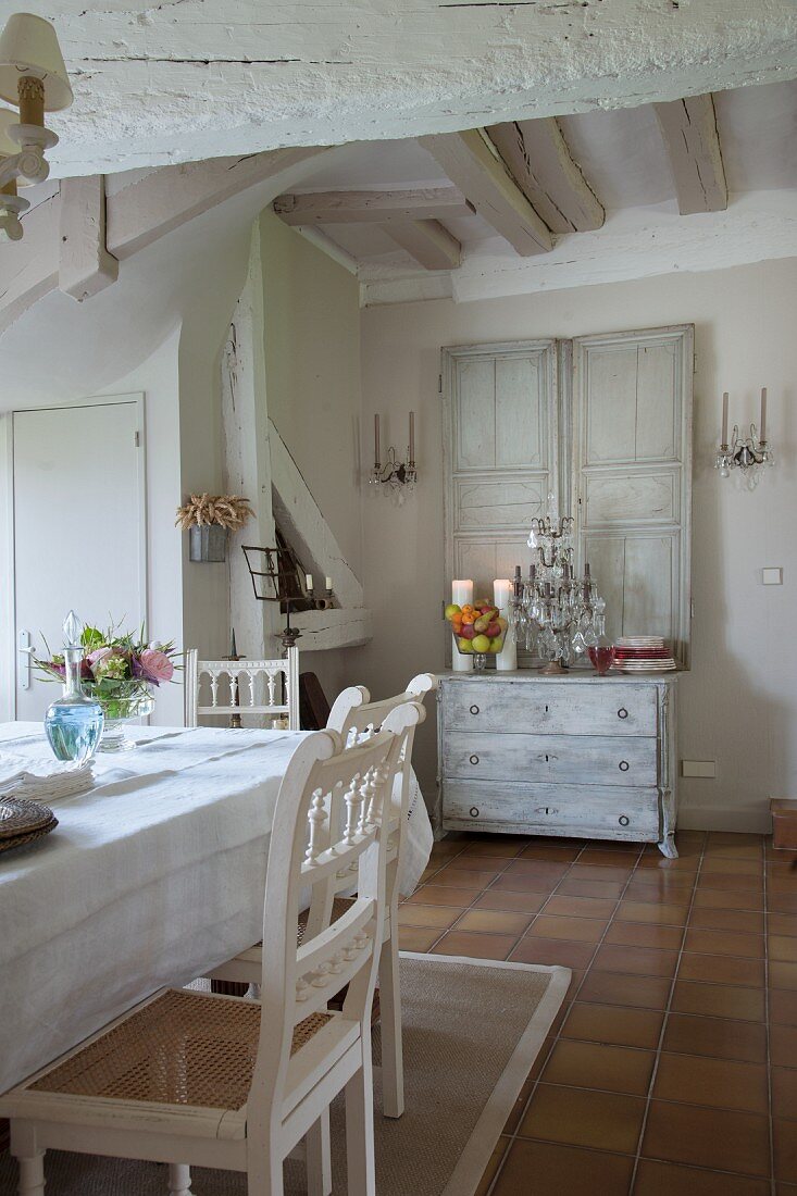 Stühle mit geschnitzten Rückenlehnen weiss lackiert an Tisch, im Hintergrund Kommode dekoriert mit Kerzen in Essbereich eines traditionellen Landhauses