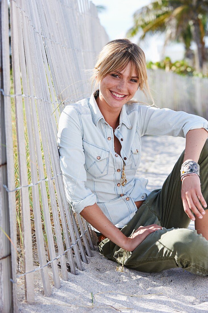 A young blonde woman sitting in the sand wearing a denim blouse and green trousers