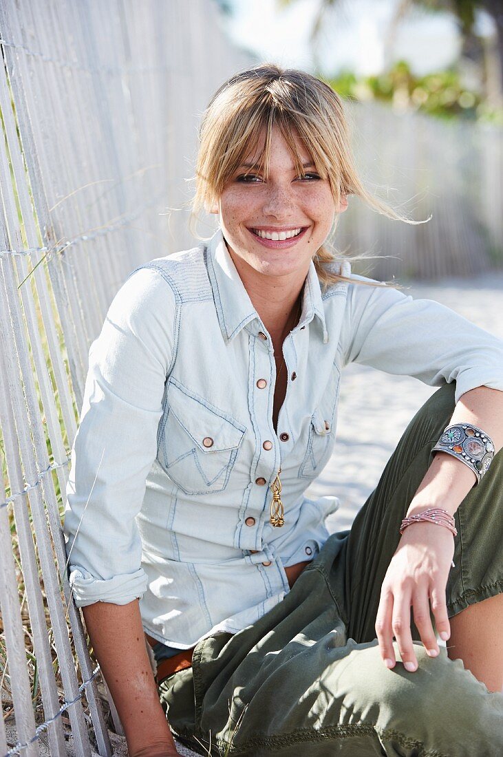 A young blonde woman sitting in the sand wearing a denim blouse and green trousers