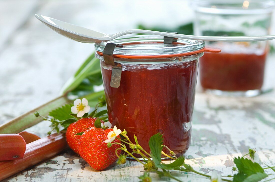 Erdbeer-Rhabarber-Marmelade in Gläsern, frische Erdbeeren, Rhabarber und Erdbeerblüten auf Holztisch