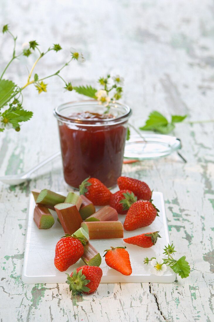 A jar of strawberry and rhubarb jam, fresh strawberries and rhubarb on a chopping board